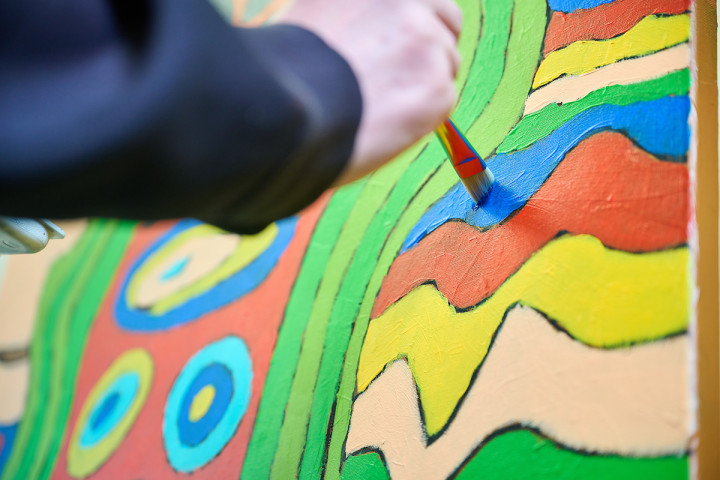 Je ziet iemands hand die met een kwast blauwe verf op een kleurrijk schilderij strijkt. Het schilderij bestaat uit golfjes, lijnen en cirkels in diverse kleuren.